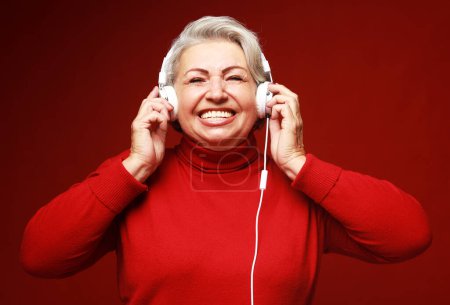 Foto de Close up photo of happy senior caucasian woman with headphones isolated over red background. Lifestyle and old people concept. - Imagen libre de derechos