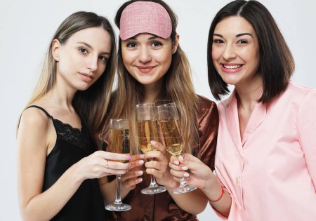 Photo for Three young happy women dressed in pajamas drink champagne and have fun, pajama party and friendship concept. - Royalty Free Image