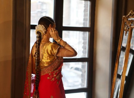 Téléchargez les photos : Female model Hindu Bride in saree, wearing gold and jasmine flower garlands in the hair near mirror, back view. - en image libre de droit