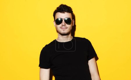 Foto de Concepto de estilo de vida, moda y gente: Retrato de un joven guapo vestido casual y con gafas de sol sobre fondo amarillo - Imagen libre de derechos
