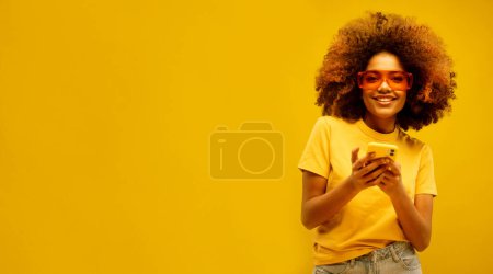 Téléchargez les photos : Style de vie, tehnology et concept de personnes : Portrait de fille afro-américaine rêveuse inspirée utiliser son smartphone, porter t-shirt jaune isolé sur fond de couleur jaune - en image libre de droit