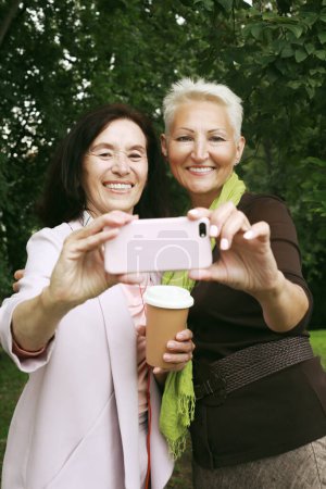 Photo for Two charming senior women friends holds a cup of coffee to go and making selfie in summer park. Lifestyle, friendship and gold age concpet. - Royalty Free Image