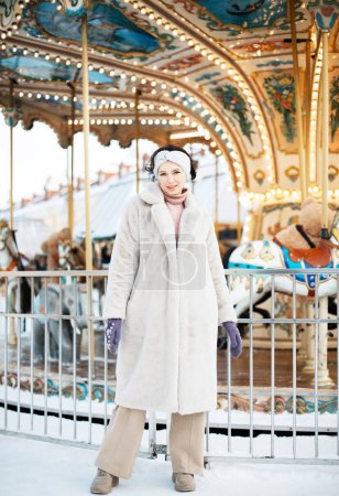 Téléchargez les photos : Une belle jeune femme brune dans un manteau en fausse fourrure et des vêtements de serpent pose à côté d'un carrousel à un marché de Noël. - en image libre de droit