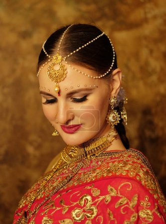 Foto de Lovely young woman wearing traditional indian red sari and jewelry, beauty smiles and looks down. Indian, Muslim, Arabic culture. - Imagen libre de derechos