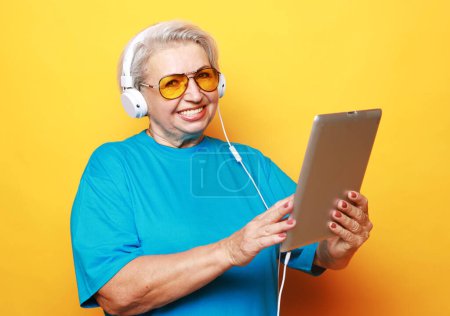 Téléchargez les photos : Education, old people and tehnology concept: Elderly woman in headphones with tablet over yellow background - en image libre de droit