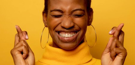 Foto de Portrait of young African American woman awaits for miracle, prays and hopes dreams come true, crosses fingers for good luck, closes eyes, smiles broadly, wears yellow jumper - Imagen libre de derechos