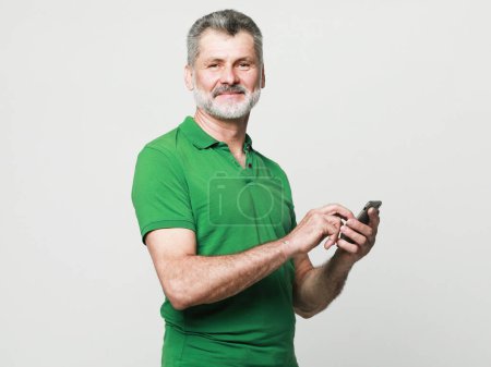 Téléchargez les photos : Smiling elderly gray-haired mustache bearded man in casual green shirt posing isolated on grey background studio. People, tehnology and lifestyle concept. Using mobile phone, typing sms message. - en image libre de droit