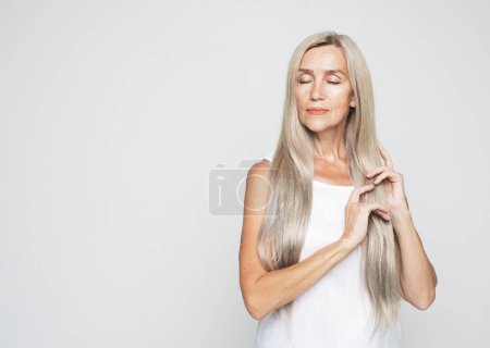 Téléchargez les photos : Concept beauté, mode et personnes âgées : belle femme âgée touche de longs beaux cheveux gris sur fond gris clair. - en image libre de droit