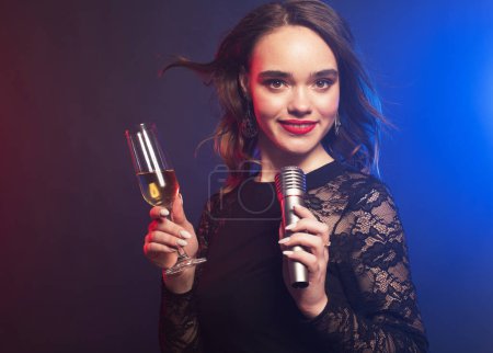 Foto de Lifestyle, party and people concept: Young pretty woman wearing black dress, holding a glass of wine and singing into microphone over blue background - Imagen libre de derechos