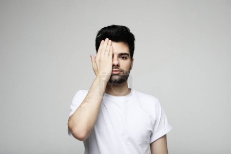 Téléchargez les photos : Young handsome brunette bearded man wearing casual white t-shirt covering one eye with hand over grey background - en image libre de droit