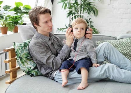 Foto de A young father holds his little son on his lap and lets him listen to music on headphones. Lifestyle and family concept. - Imagen libre de derechos