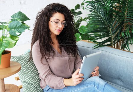Foto de Estilo de vida, tehnología, educación y concepto de personas. Joven afroamericana hembra está utilizando una tableta digital mientras está sentado en el sofá. - Imagen libre de derechos