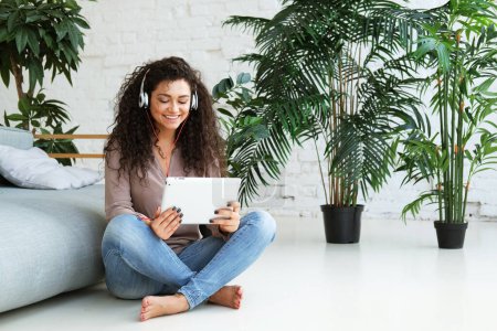 Photo for People, technology and leisure concept - happy african american young woman sitting on floor near sofa with tablet pc computer and headphones listening to music at home - Royalty Free Image