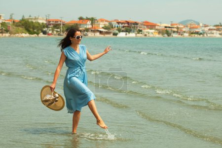 Foto de Mujer morena joven en un vestido azul caminando descalza en una playa y cuelga sus pies en el agua. Felices vacaciones de verano. - Imagen libre de derechos