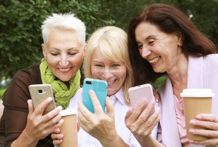 Téléchargez les photos : De charmantes vieilles femmes rient joyeusement et regardent les écrans de leurs smartphones. S'amuser à la retraite un après-midi d'été. Âge d'or. - en image libre de droit