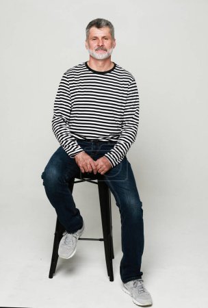 Téléchargez les photos : Elderly bearded man in a striped cardigan sits on a chair on a gray studio background. Lifestyle, emotion and old people concept. - en image libre de droit