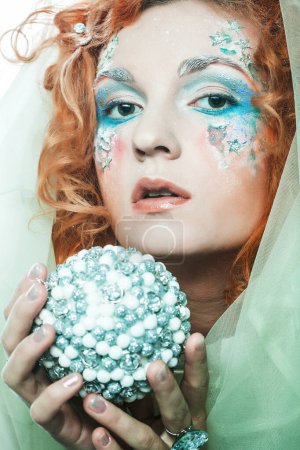 Party, christmas and people concept: Beautiful redhead woman in christmas party costume holding christmas ball. Creative makeup, hairstyle, close-up portrait. Winter style, snow queen.