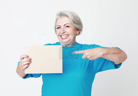 Photo for A happy and charming elderly woman holds a blank sheet of paper in her hands and points at it with her finger. Isolated over white background. - Royalty Free Image