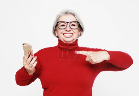Foto de Lifestyle, technology and old people concept: A picture of mature woman wearing red sweater with new smartphone. She has tested it and admitted this phone is a good one. - Imagen libre de derechos