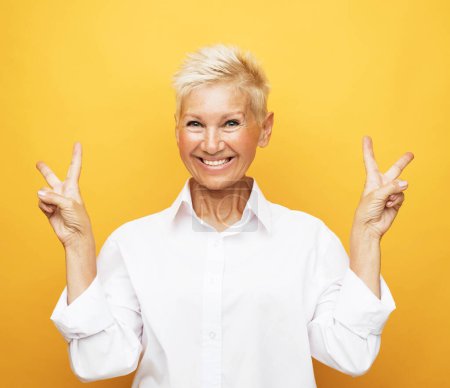 Téléchargez les photos : Portrait of happy and charming european senior woman with blond short hair laughing and show v-sign over yellow background - en image libre de droit