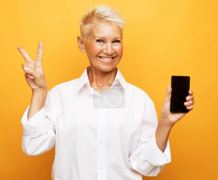 Téléchargez les photos : Modern lifestyle, old people, communication and electronic gadgets concept. Attractive elderly woman with pixie blonde hair presents new smartphone over yellow background. - en image libre de droit