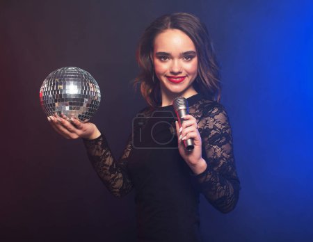 Photo for Lifestyle, party and people concept: young woman wearing black dress, holding disco ball and singing into microphone over dark background - Royalty Free Image