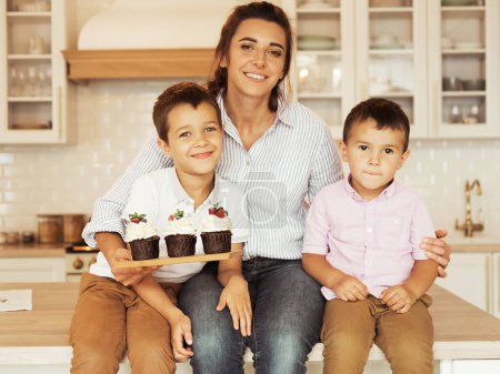 Foto de Familia feliz en la cocina. Mamá y dos niños pequeños, hermanos, están comiendo pastelitos caseros. Creatividad familiar, aficiones, actividades conjuntas con niños. - Imagen libre de derechos