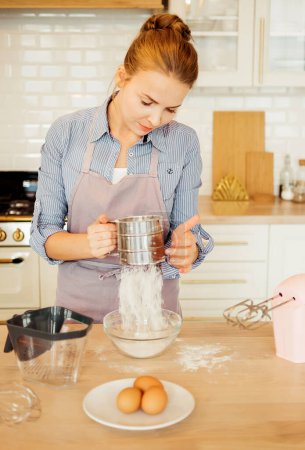Photo for A young woman sifts flour through a sieve, for dough, baking, cake, pie. Modern kitchen. Hobby. - Royalty Free Image