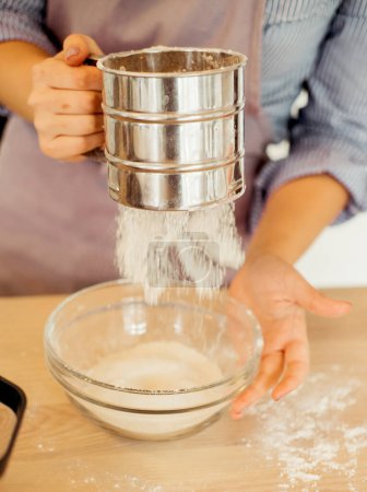 Foto de A young woman sifts flour through a sieve, for dough, baking, cake, pie. Modern kitchen. Close up. - Imagen libre de derechos
