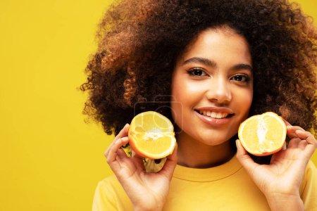 Foto de Estilo de vida, comida, dieta y concepto de personas: Foto de una joven afroamericana que sostiene rebanadas de naranja se ven sorprendidas aisladas sobre un fondo de color amarillo. - Imagen libre de derechos
