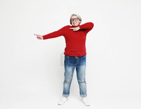 Téléchargez les photos : Smiling elderly gray-haired blonde woman lady 60s years old in red casual sweater pointing index finger aside up on mock up copy space isolated on white background studio portrait - en image libre de droit