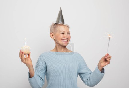 Photo for Happy middle aged female wearing conical hat celebrating birthday with cupcake with candle and sparkler over grey background - Royalty Free Image