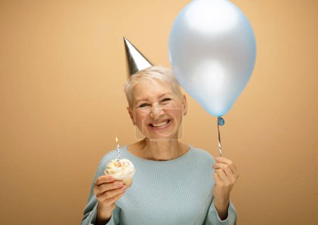Photo for People, party, aging and maturity concept. Beauitful elderly female wearing conical hat celebrating birthday, posing isolated with cupcake with candle and blue balloon in her hands. Happy time. - Royalty Free Image