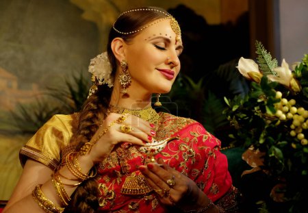 Téléchargez les photos : Charming Indian bride dressed in Hindu red traditional wedding clothes embroidered with gold sitting on sofa near flowers, smiles and holding small gold box. - en image libre de droit
