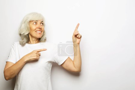 Foto de Retrato de mujer perfecta, agradable, vieja, impresionada en camiseta blanca demostrar, mirando, señalando con los dedos hacia arriba para copiar el espacio, con la boca abierta, de pie sobre fondo gris claro - Imagen libre de derechos