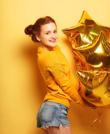 Téléchargez les photos : Gros plan de mignonne fille rouge debout dans un studio, souriant largement et jouant avec des ballons d'or. Fond jaune
. - en image libre de droit