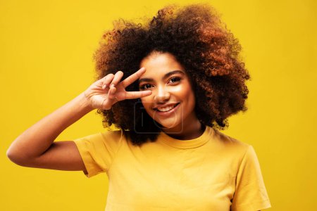 Foto de Lifestyle, emotion and people concept: Charming fashionable young black curly woman 20s wears yellow shirt showing covering eye with victory sign isolated on yellow background studio portrait - Imagen libre de derechos