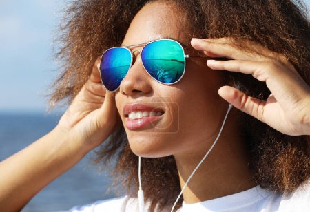 Photo for Charming amazing afro american young woman in sunglasses listening to music in headphones on her mobile phone. Dressed casual. Fun time outdoors near sea. Lifestyle. - Royalty Free Image