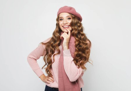 Foto de Smiling blonde female model looks at the camera. Young woman with curly hair wearing a pink sweater, scarf, and beret over grey background. - Imagen libre de derechos