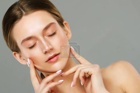 Foto de Beautiful young woman with closed eyes and clean fresh skin touching her face over grey background. Cosmetology , beauty and spa concept. - Imagen libre de derechos