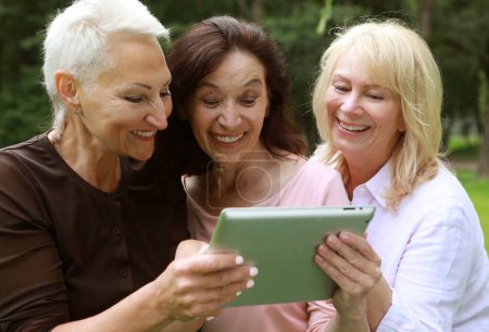 Photo for Three elderly women are smiling and looking news at the at screen of the tablet in the park on a summer day. The concept of friendship, happy old age and emotions. - Royalty Free Image