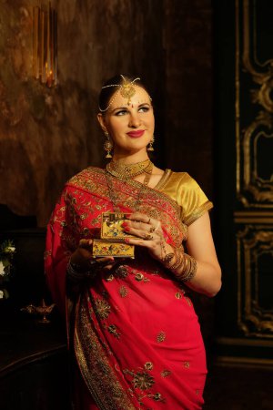 Foto de Charming young woman in traditional indian red sari and jewelry holding golden casket, beautiful woman smiling. Indian, Muslim, Arabic culture. - Imagen libre de derechos