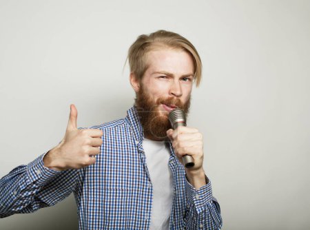 Téléchargez les photos : Young redhair bearded man wearing a blue shirt and glasses holding a microphone and singing over grey background. - en image libre de droit