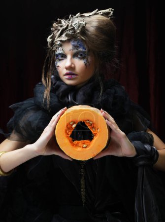 Photo for Young beautiful woman in a witch costume with bright makeup and hairstyle holding a pumpkin, a symbol of halloween. Party. Holiday. Carnival. - Royalty Free Image