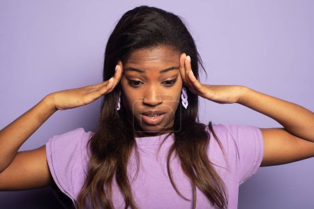 Photo for Young afro american woman clutching her head with her palms while suffering from a migraine. Portrait on a purple color background. - Royalty Free Image