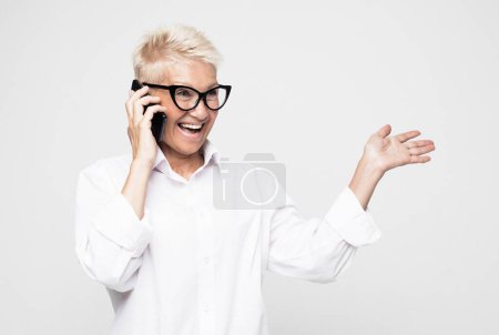 Foto de Lifestyle, technology and old people concept: Great news. Happy senior woman using mobile phone over grey background. Beautiful stylish elderly lady talking on cellphone with happy emotions. - Imagen libre de derechos