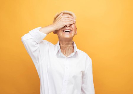Foto de Laughing mature woman 60 years old with short hair closes her eyes with her palm on a yellow background - Imagen libre de derechos