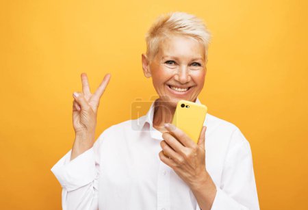 Téléchargez les photos : Modern lifestyle, old people, communication and electronic gadgets concept. Attractive elderly woman with pixie blonde hair presents new smartphone over yellow background. - en image libre de droit