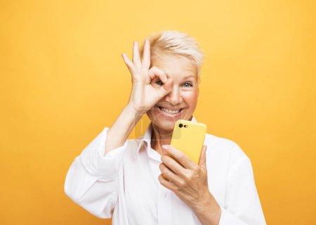 Téléchargez les photos : Happy mature senior woman with short hair holding smartphone using mobile and show finger ring ok-sign over yellow background. - en image libre de droit