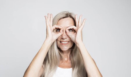 Foto de Estilo de vida, emoción y el concepto de las personas mayores: Mujer de pelo gris de edad con camisa blanca haciendo gesto bien sorprendido con la cara sorprendida, los ojos mirando a través de los dedos. expresión incrédula. - Imagen libre de derechos